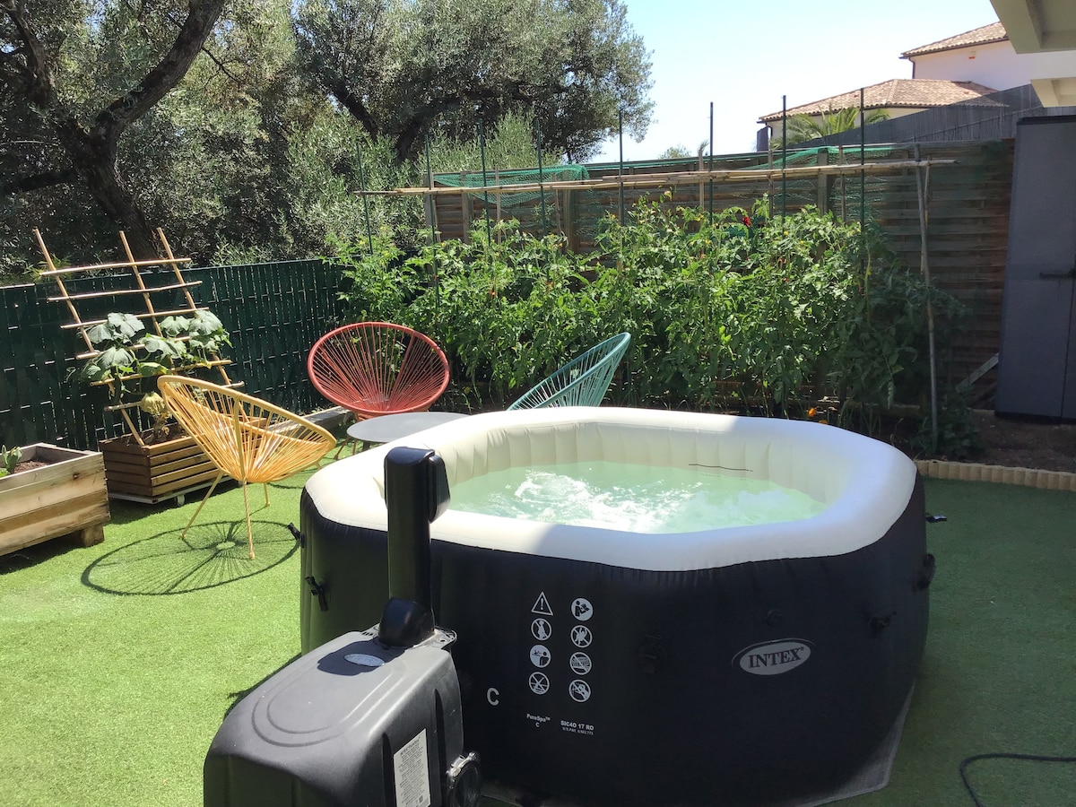 A garden area features a portable inflatable jacuzzi, surrounded by greenery and vegetable plants. Colorful lounge chairs in turquoise and yellow invite relaxation. The space is set on faux grass, creating a comfortable and inviting outdoor atmosphere.