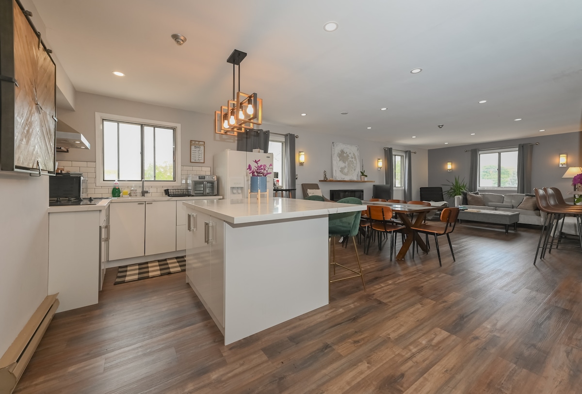 An open kitchen is accompanied by a dining area and living space, featuring a large island with seating. Natural light enters through multiple windows, illuminating the wood flooring. Dining chairs surround a modern table, while a comfortable seating arrangement is positioned nearby.