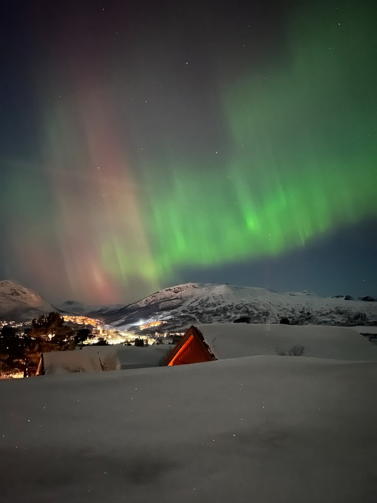 The night sky is illuminated by vibrant green and purple auroras, casting an ethereal glow over the snowy landscape. Below, rooftops emerge from the white blanket, while distant mountain peaks create a stunning backdrop to the captivating natural display.