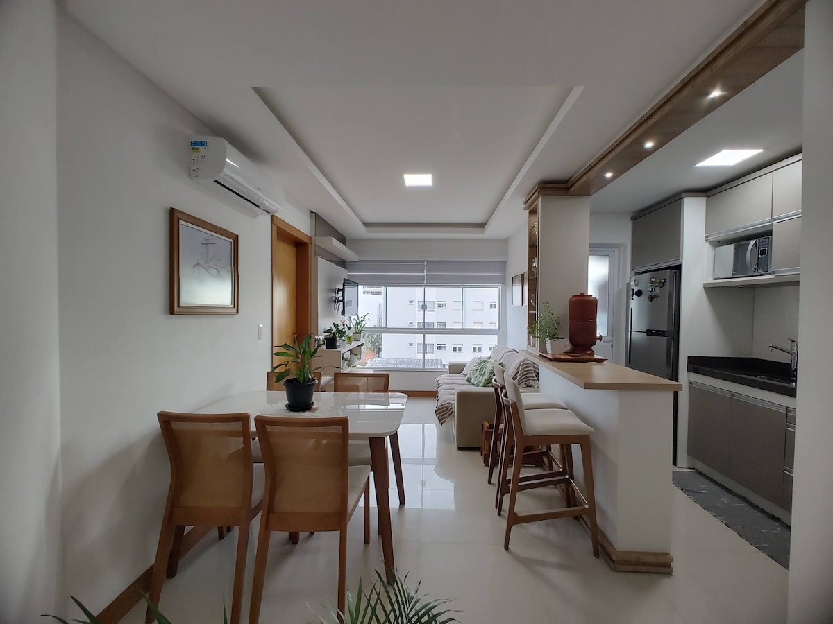 A bright and open plan living area features a dining table with four chairs, allowing for comfortable seating. A sleek kitchen is visible in the background, equipped with modern appliances, while large windows bring in natural light, enhancing the overall spaciousness.