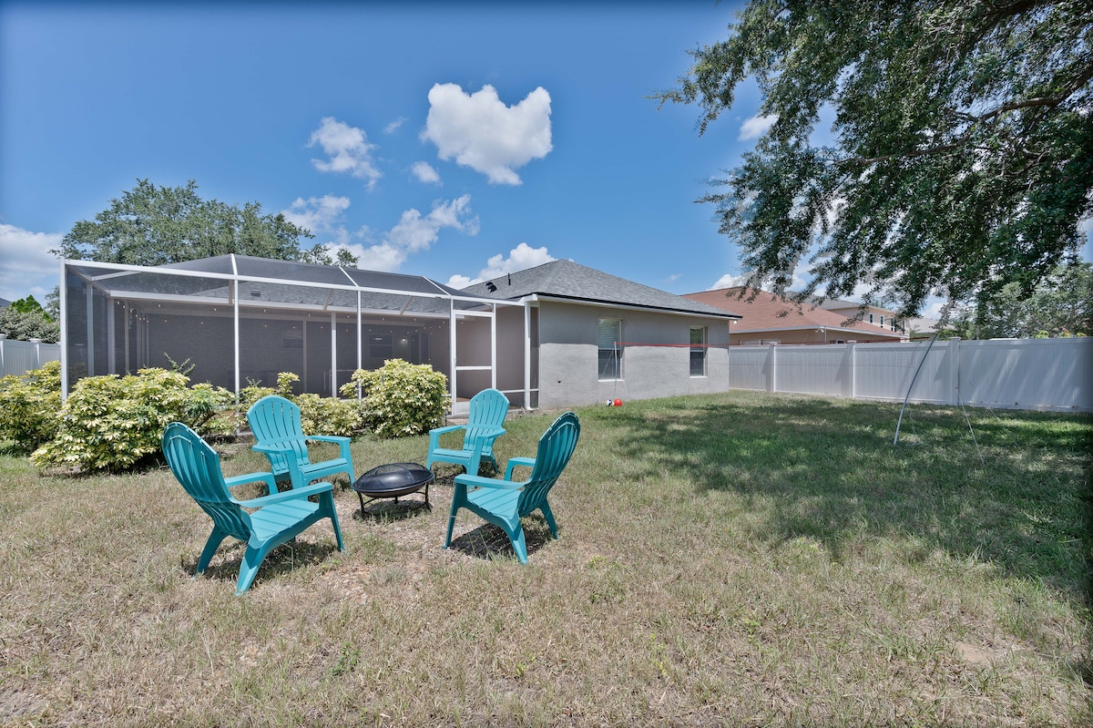 A spacious backyard is shown, featuring a grassy area with four turquoise adirondack chairs arranged around a fire pit. A screened patio area is visible in the background, alongside a few shrubs and trees. A fence encloses the yard, offering privacy.