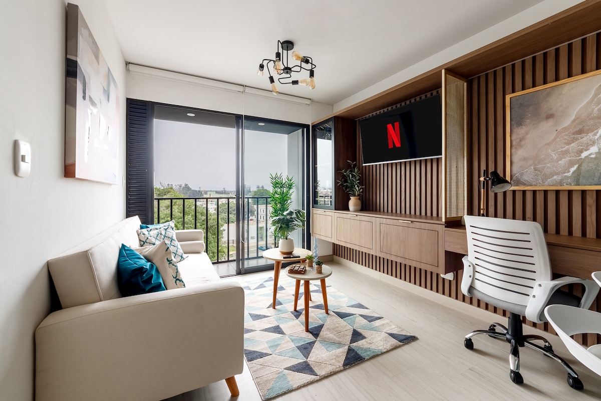 A bright living area showcases a modern design, featuring a sofa adorned with decorative pillows. A geometric area rug is placed under a coffee table, which holds small plants. A wall-mounted SmartTV displays the Netflix logo, and a workspace is positioned in the corner near large glass doors leading to a balcony.