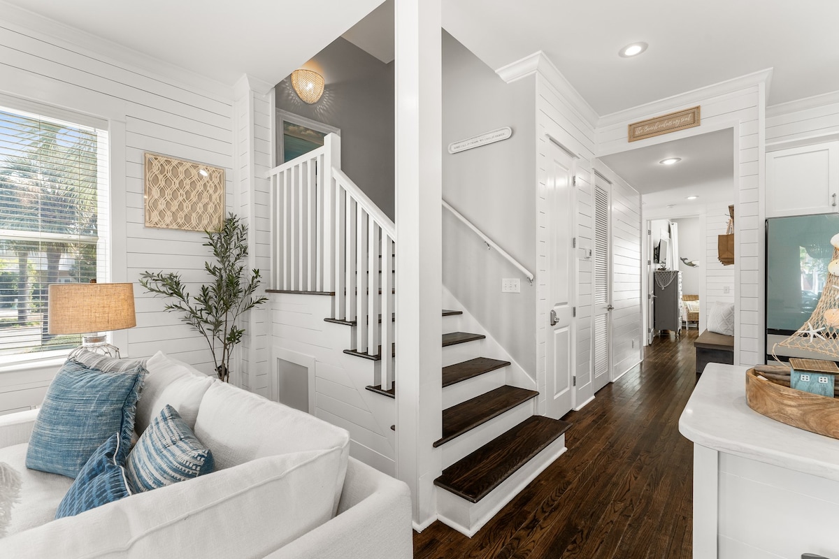 A bright entryway features light wood paneling and a cozy white sofa adorned with blue accent pillows. A staircase with dark wooden steps leads to the upper level, while a decorative light and greenery add a touch of warmth to the space.