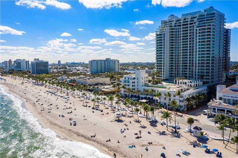 An aerial view captures the sandy Ft. Lauderdale Beach lined with sunbathers and beach chairs. The shoreline transitions into vibrant cityscape, with tall buildings in the background. Waves gently lap at the beach, under a bright blue sky with scattered clouds.