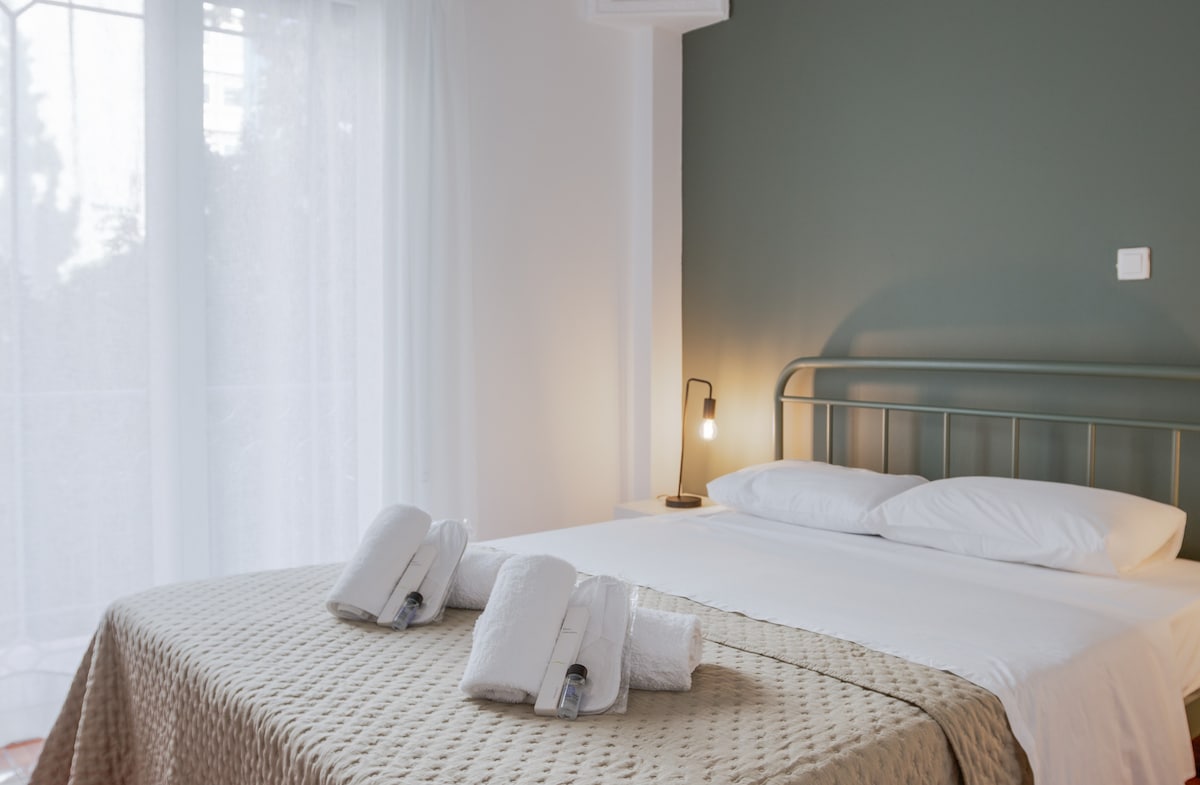 Მყუდრო საძინებელი რბილი კრემისფერი საწოლის გადასაფარებლით, მწვანე კედლით და თანამედროვე განათებით. Ახალი პირსახოცები სუფთად არის მოწყობილი საწოლზე, რაც სტუმართმოყვარე და დასასვენებელ ატმოსფეროს ქმნის. Ბუნებრივი სინათლე ფანჯრიდან იღვრება და სიმშვიდის ატმოსფეროს აუმჯობესებს.