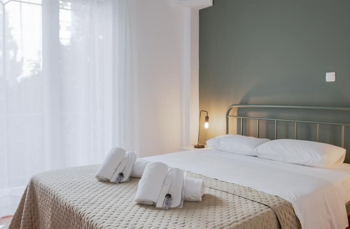 Útulná spálňa s mäkkou béžovou posteľnou prikrývkou, zelenou dekoratívnou stenou a moderným osvetlením. Čerstvé uteráky sú úhľadne usporiadané na posteli a vytvárajú príjemnú a relaxačnú atmosféru. Prirodzené svetlo prúdi cez okno a zvyšuje pokojnú atmosféru.