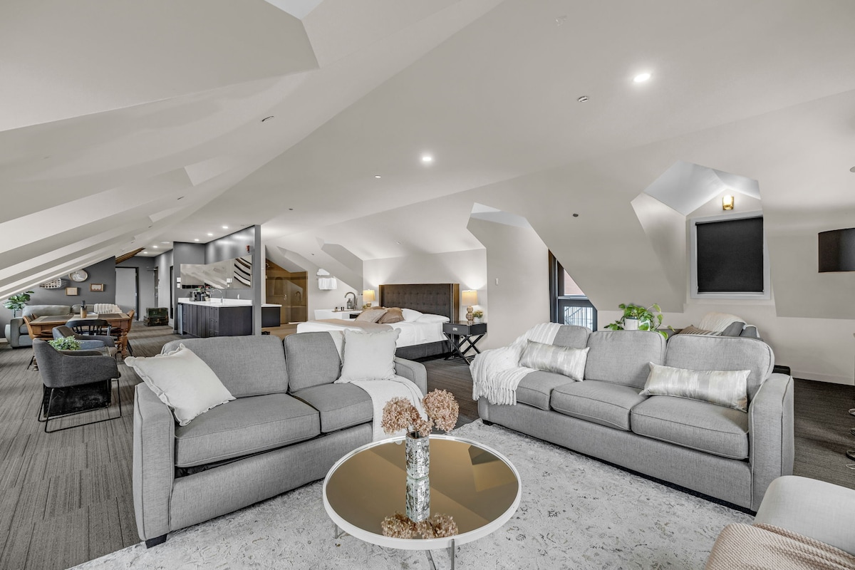 An open-concept living area features two spacious gray sofas arranged around a circular coffee table. A dining area can be seen in the background, along with a well-equipped kitchenette. Natural light filters in through the angular ceilings, highlighting the room's modern design.