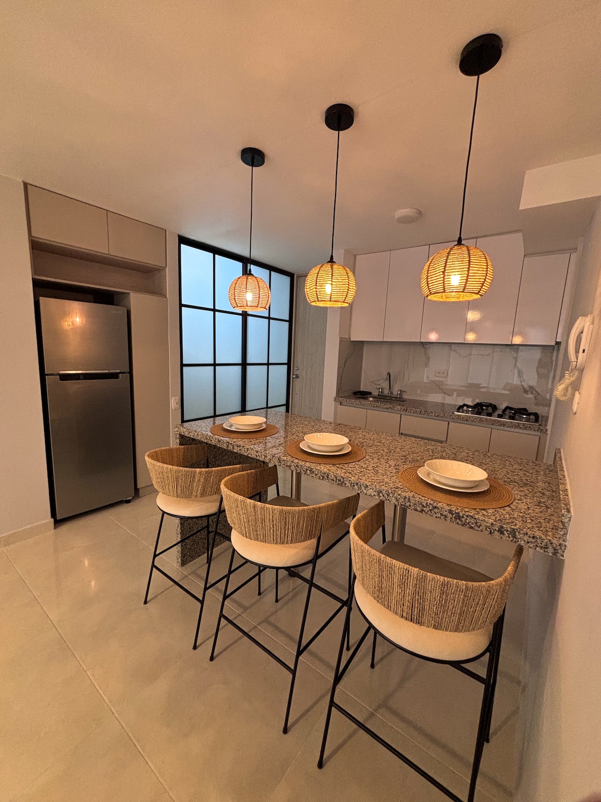 A modern kitchen space features a granite countertop with four bar stools. Pendant lighting hangs above, illuminating the area. The equipped kitchen includes cabinetry and appliances, with a glass partition allowing natural light to flow in from the adjacent space.