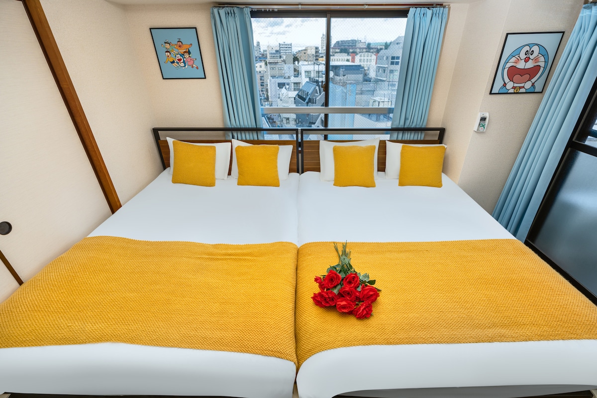 A bright sleeping area features two spacious beds, each adorned with yellow throw blankets and several decorative pillows. A vase with red roses is placed on the beds, while large windows allow natural light to brighten the room and offer urban views.