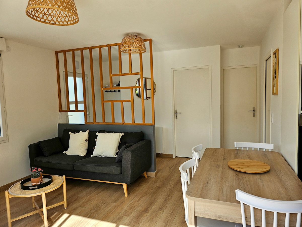 The living area features a cozy sofa adorned with decorative pillows, complemented by a wooden coffee table. A dining table with four chairs is positioned nearby. Light streams through windows, highlighting the open space, which is separated by a decorative wooden partition.