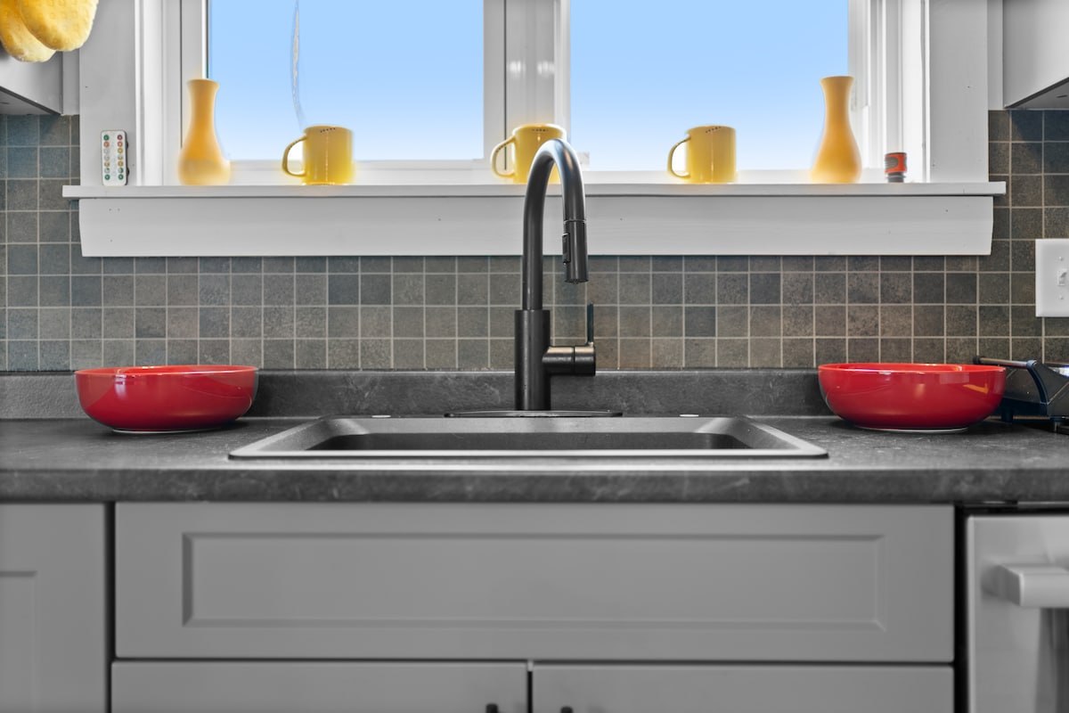 The kitchen sink area features a modern faucet above a stainless steel basin. Two vibrant red bowls are placed on the countertop, while cheerful yellow mugs are arranged along the window ledge, complemented by a light tile backsplash.