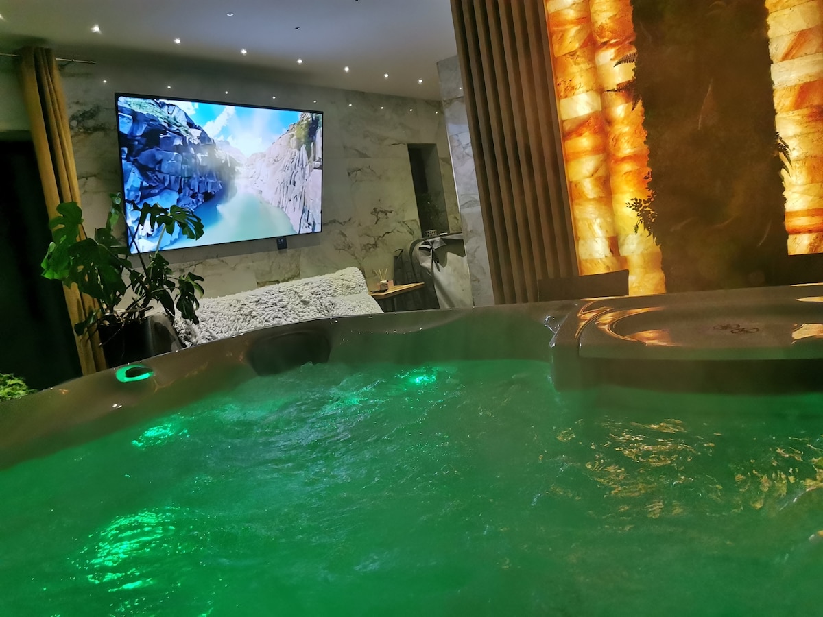 A hot tub with bubbling water is positioned in a stylish indoor space. Soft green lighting reflects off the water's surface. A large television screen is mounted on the wall, displaying a scenic nature image. Surrounding plants add a touch of greenery to the environment.