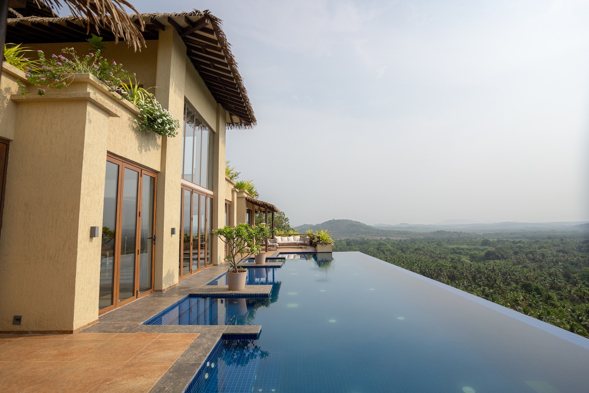 An infinity pool extends from the villa, offering sweeping views of the surrounding landscape. The pool area is adorned with potted plants and features clear blue water that reflects the sky. Expansive glass doors lead into the villa, enhancing the scenic connection.