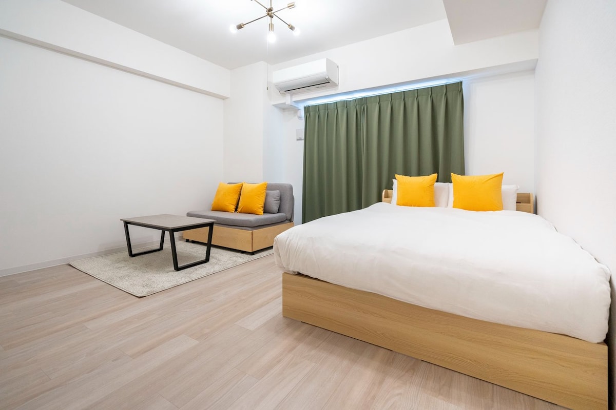 A spacious room features a comfortable bed with white linens and bright orange accent pillows. A gray sofa sits opposite a low coffee table on a soft rug. Natural light enters through a large window covered by green curtains, contributing to a calm and inviting environment.