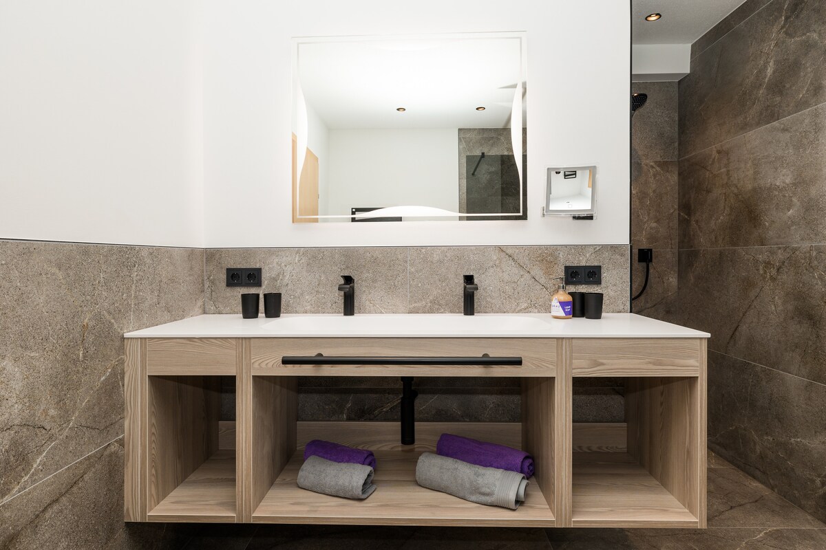 A modern bathroom features a wooden vanity with an elegant sink and a large mirror above. The space is complemented by natural stone walls and well-placed lighting. Towels are neatly arranged on the shelves, enhancing a clean and organized appearance.