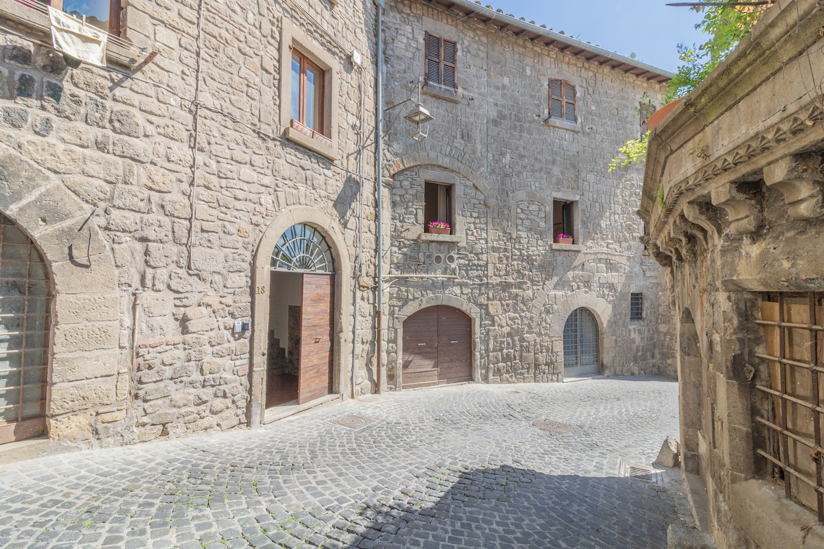 A cobblestone street is lined with historic stone buildings, featuring arched doorways and wooden windows adorned with flower boxes. Soft light illuminates the area, revealing the building textures and architectural details, creating an inviting scene in the heart of a medieval setting.