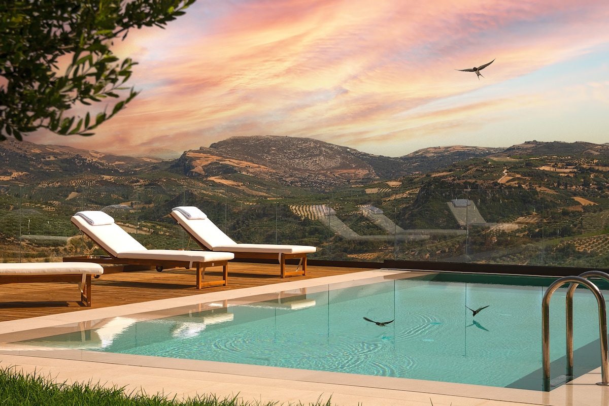 A serene pool area is presented with two lounge chairs positioned beside the water. The backdrop features a landscape of rolling hills adorned with vineyards and olive groves, while a vibrant sky adds warmth to the scene.