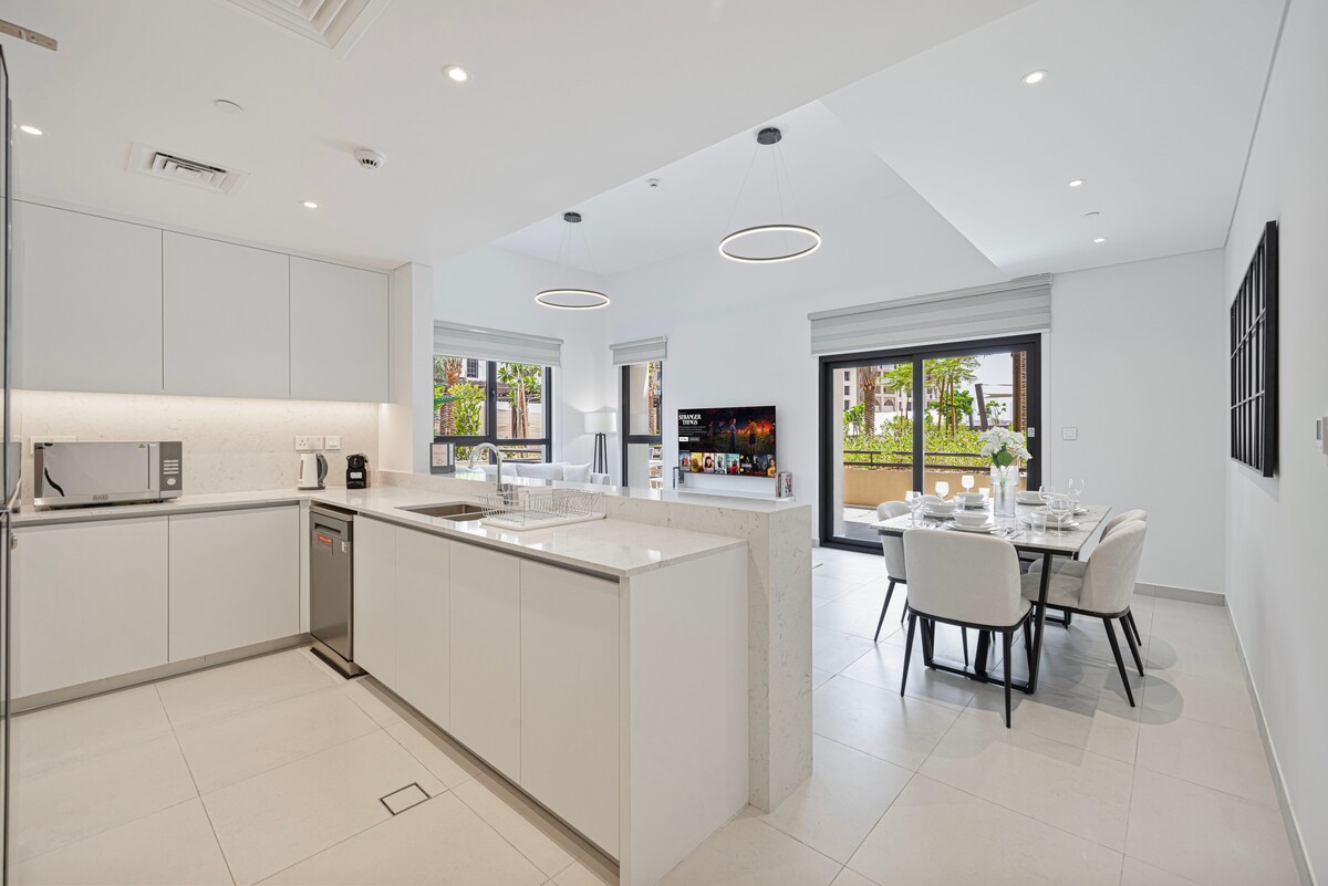 The modern kitchen features sleek cabinetry and a spacious island with seating. A dining table for four is positioned nearby, complemented by large windows that provide natural light and views of the outdoor terrace. A smart TV adds a contemporary touch to the living area.