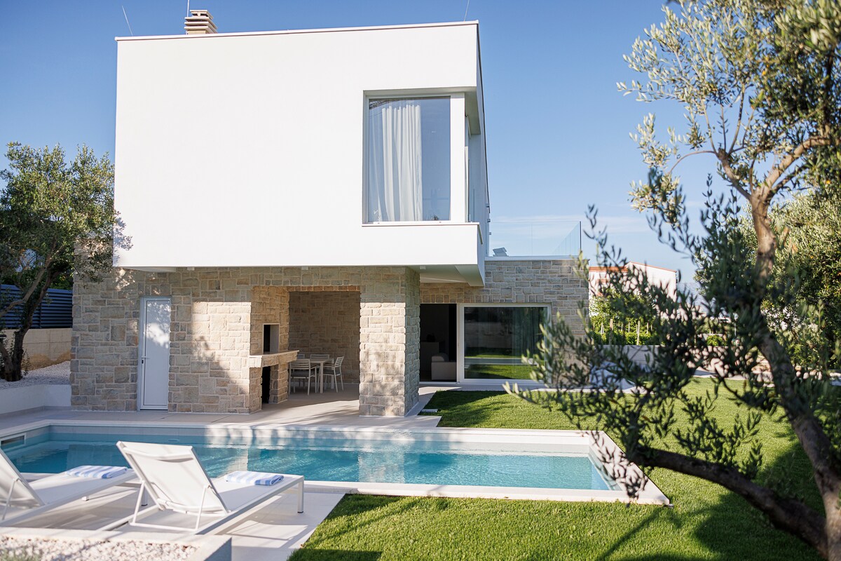 The modern villa features a sleek architectural design with a white exterior and natural stone accents. A pool is set in front of the building, surrounded by lounge chairs. Lush green grass is visible around the pool, enhancing the outdoor relaxation area.