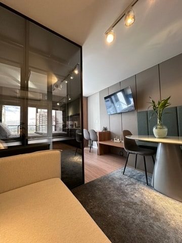 A modern living space features a cozy sofa facing a glass partition that separates a sleeping area. A table with comfortable chairs is positioned nearby, complemented by a television mounted on the wall. Natural light filters through large windows, enhancing the overall ambiance.