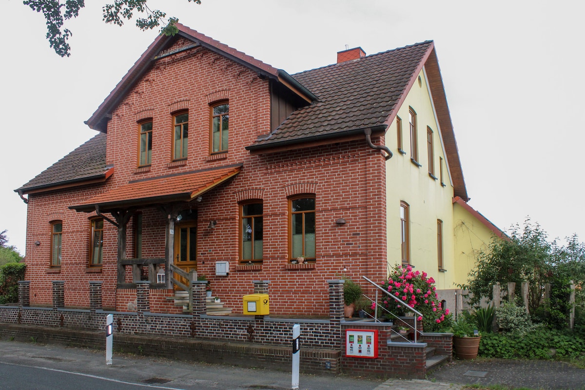 The exterior of a historic building is displayed, featuring a brick facade with a combination of red and pale yellow colors. Large windows are visible on both stories, complemented by a covered entrance with a wooden railing. Lush greenery and potted plants are located along the pathway.