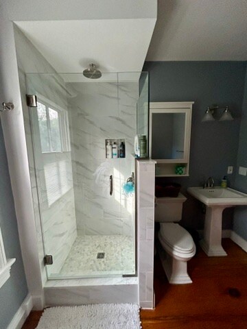 A modern bathroom is shown with a glass-enclosed shower featuring marble-like tiles and a stone floor. A pedestal sink and toilet are positioned beside the shower. Natural light enters through a window, illuminating the clean lines and neutral color scheme.