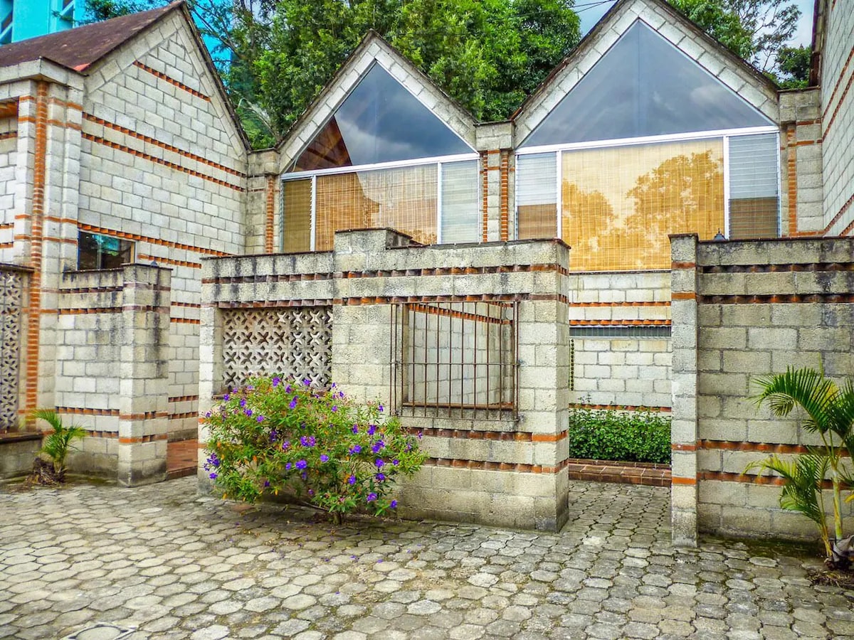 The image features a modern, two-story building with large windows reflecting the surrounding greenery. An inviting courtyard is visible, paved with stone, flanked by well-maintained flower beds and small palm plants. The textured wall design provides a unique architectural detail.