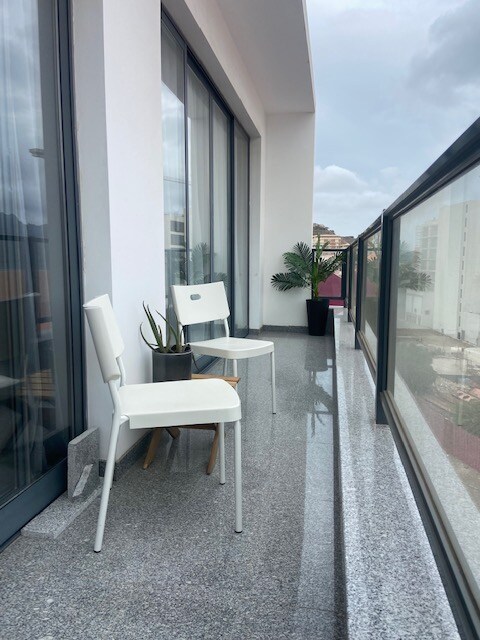 A slender balcony is adorned with two white chairs, positioned thoughtfully along the glass railing. A potted plant adds a touch of greenery, while the smooth, grey flooring reflects natural light. City views can be glimpsed beyond the railing.