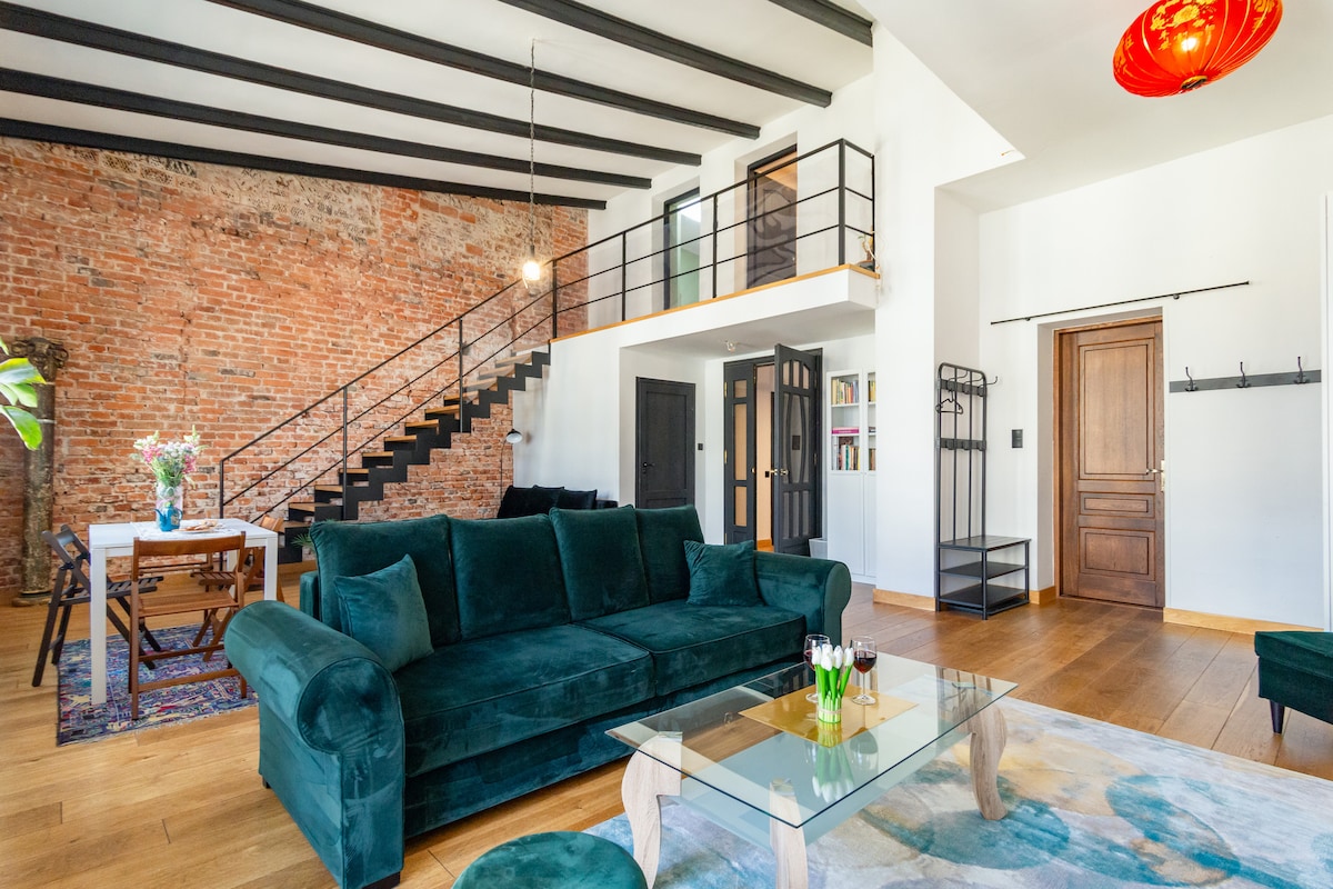 A spacious living area showcases a deep green sofa paired with a glass coffee table. Exposed brick walls add character, while wooden flooring enhances warmth. A staircase with a modern railing leads to an upper level, complemented by natural light streaming through large windows.