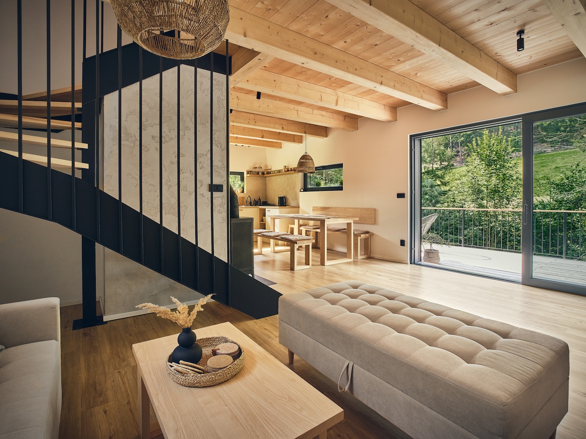 An open-plan living area features a staircase leading to the upper level, alongside a light-colored sofa and a coffee table. The dining space, visible in the background, contains a wooden table surrounded by chairs. Large glass doors provide a view of the surrounding greenery.