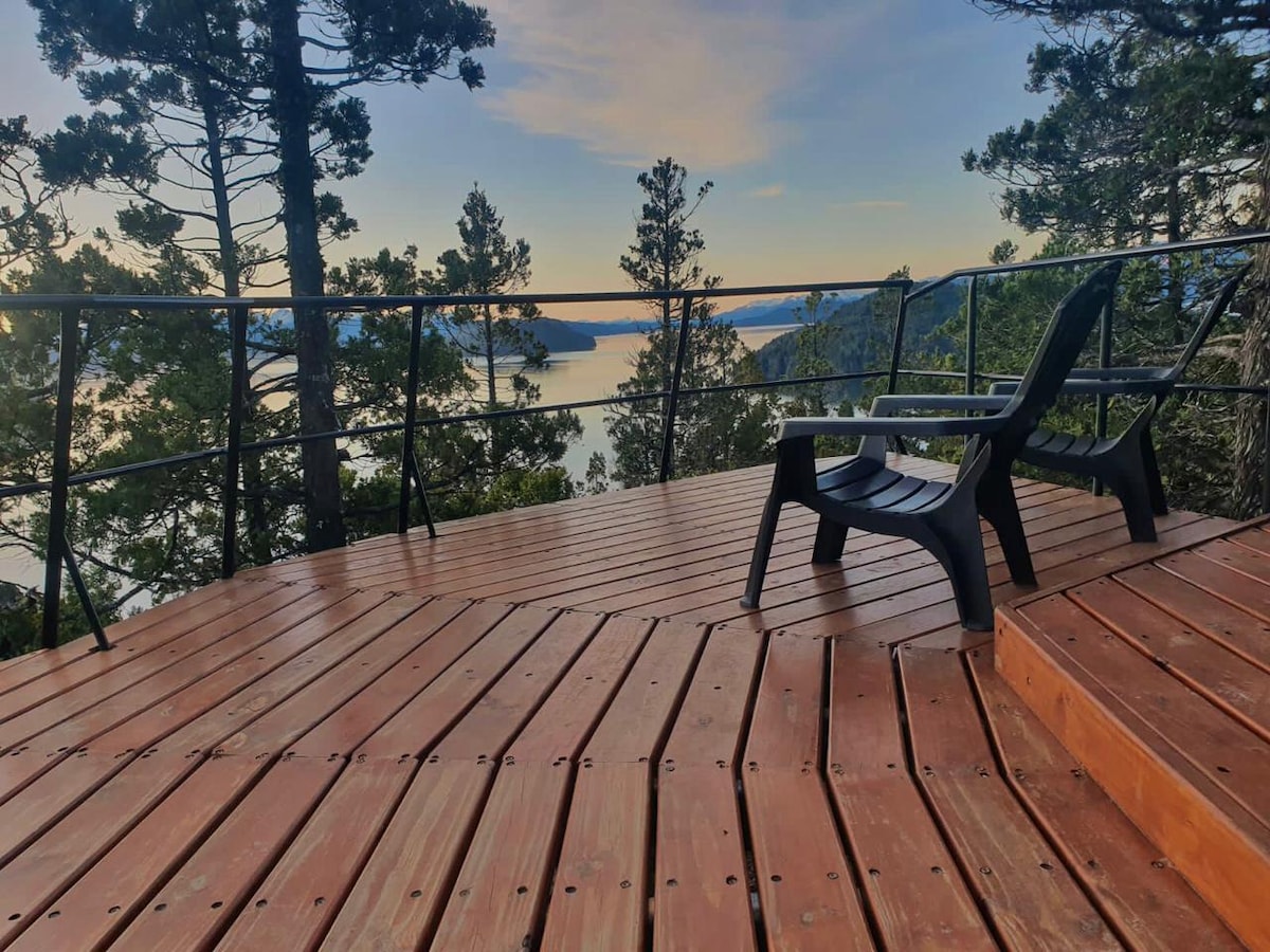 A wooden deck is presented with a spacious layout, featuring two black lounge chairs positioned for optimal views. Surrounding trees provide natural shade, while the tranquil water of the lake is visible in the background, reflecting the soft colors of the sky.