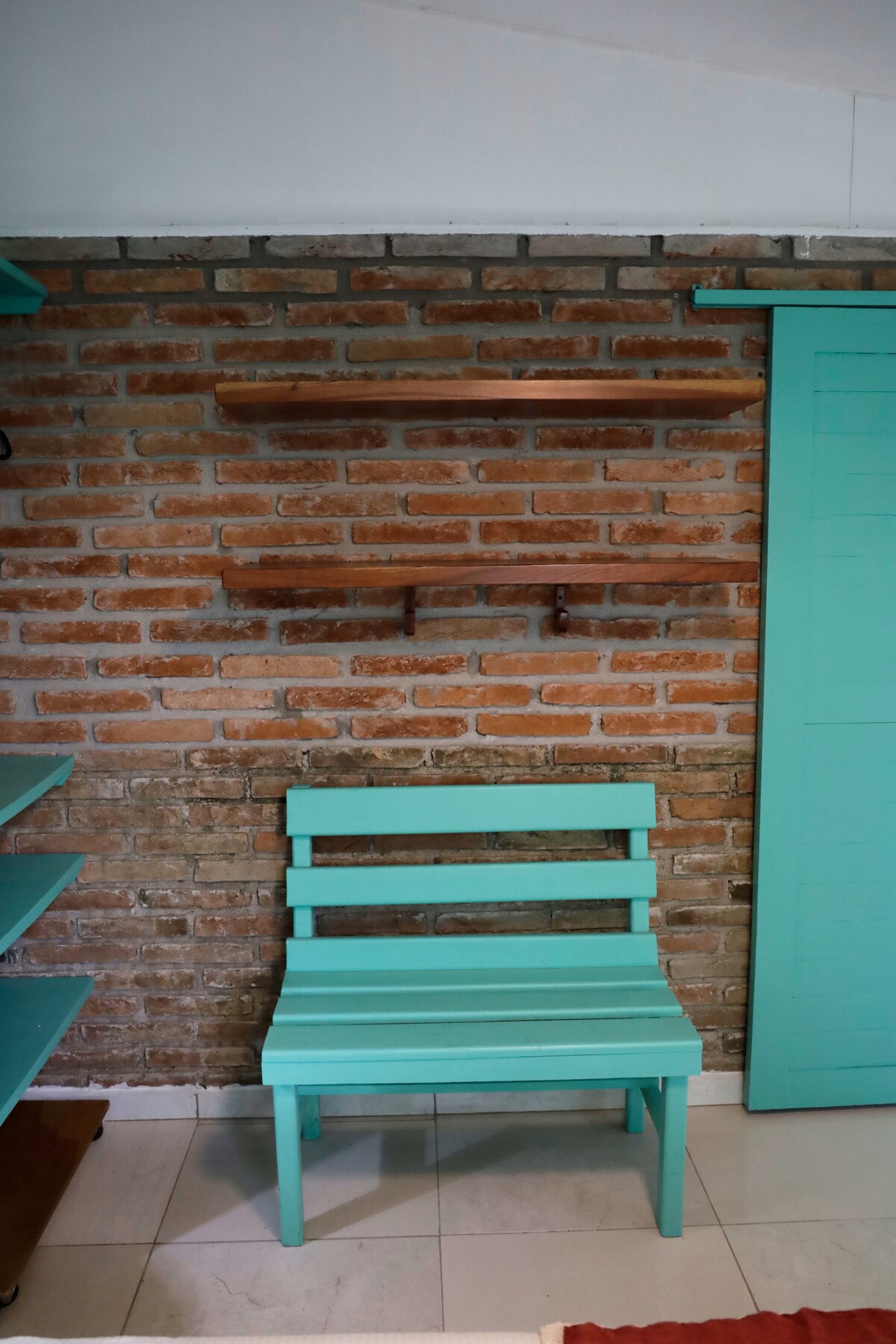 A turquoise wooden chair is positioned against a textured brick wall. Above the chair, two wooden shelves are mounted, providing a space for potential decorative items. The floor features light tiles, complementing the vibrant chair and the rustic wall.