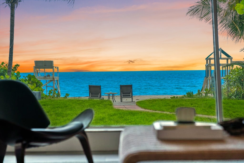 A serene view of the Atlantic Ocean is captured from inside the villa, framed by lush greenery. Two lounge chairs sit invitingly on the sand, with a wooden play structure visible in the background. The sunset casts warm hues across the water, enhancing the tranquil setting.