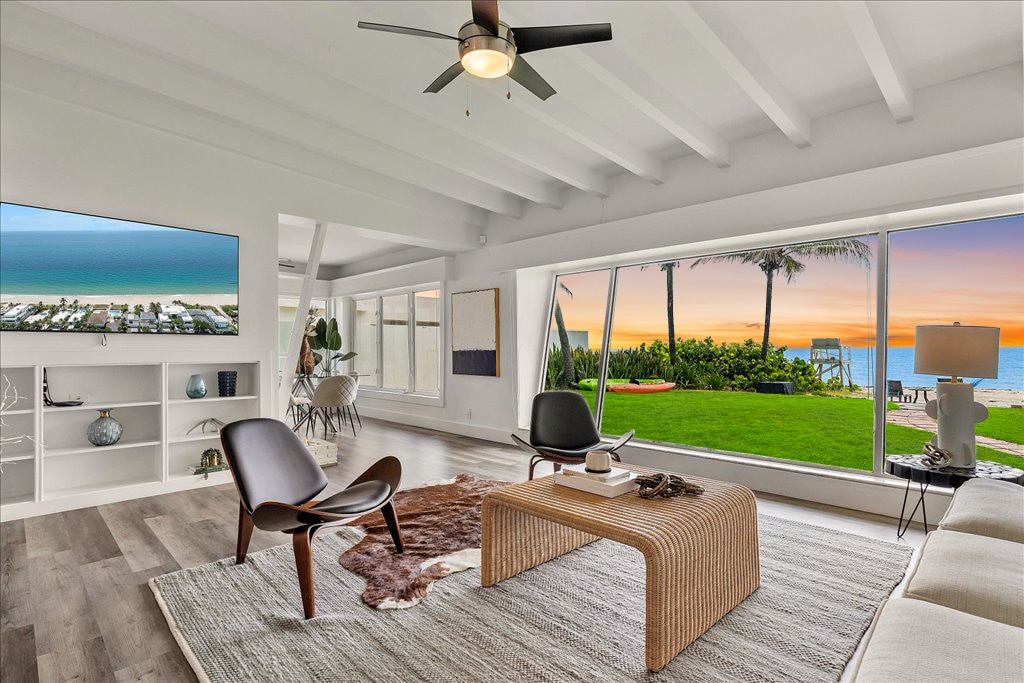 A bright and airy living room features large glass windows offering views of the ocean and sunset. The room is furnished with stylish seating and a simple coffee table atop a soft area rug. A mounted TV is positioned above a modern entertainment unit.
