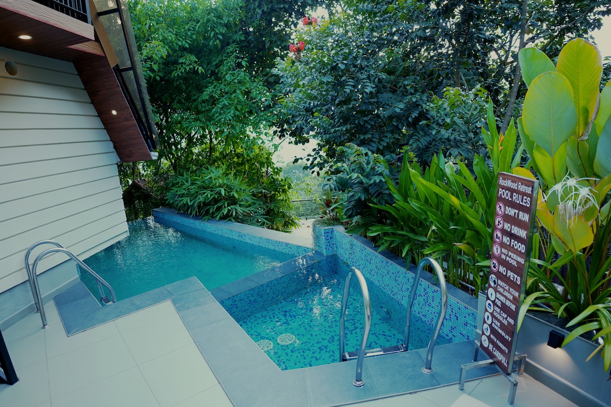 A tranquil pool area is surrounded by lush greenery, featuring a modern design with mosaic tiles and a waterfall feature. Pool rules are displayed on a sign nearby. Access to the pool is provided by stainless steel handrails.