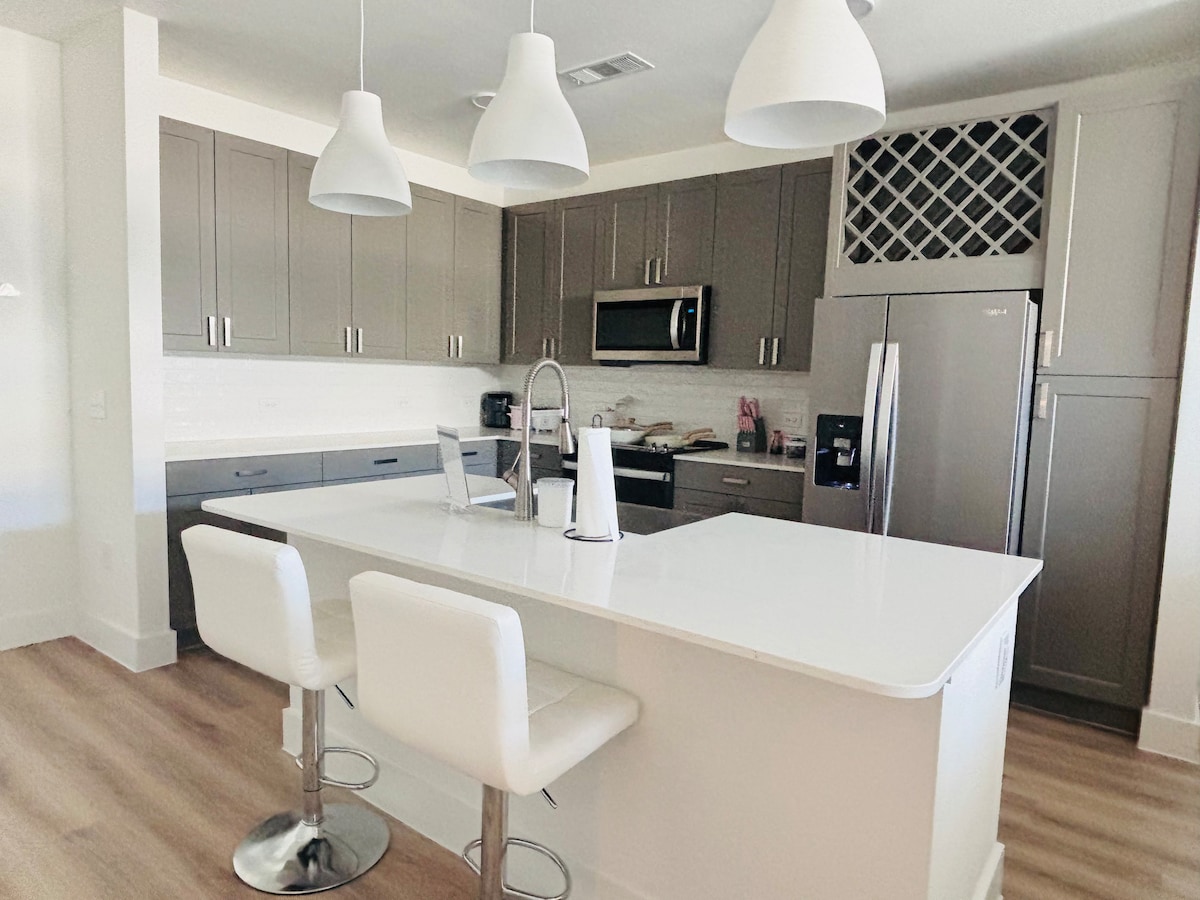 A modern kitchen design features sleek gray cabinetry complemented by white countertops. Three pendant lights hang above a spacious island with seating. Stainless steel appliances are integrated into the layout, while additional storage is provided by a wine rack above the counter.