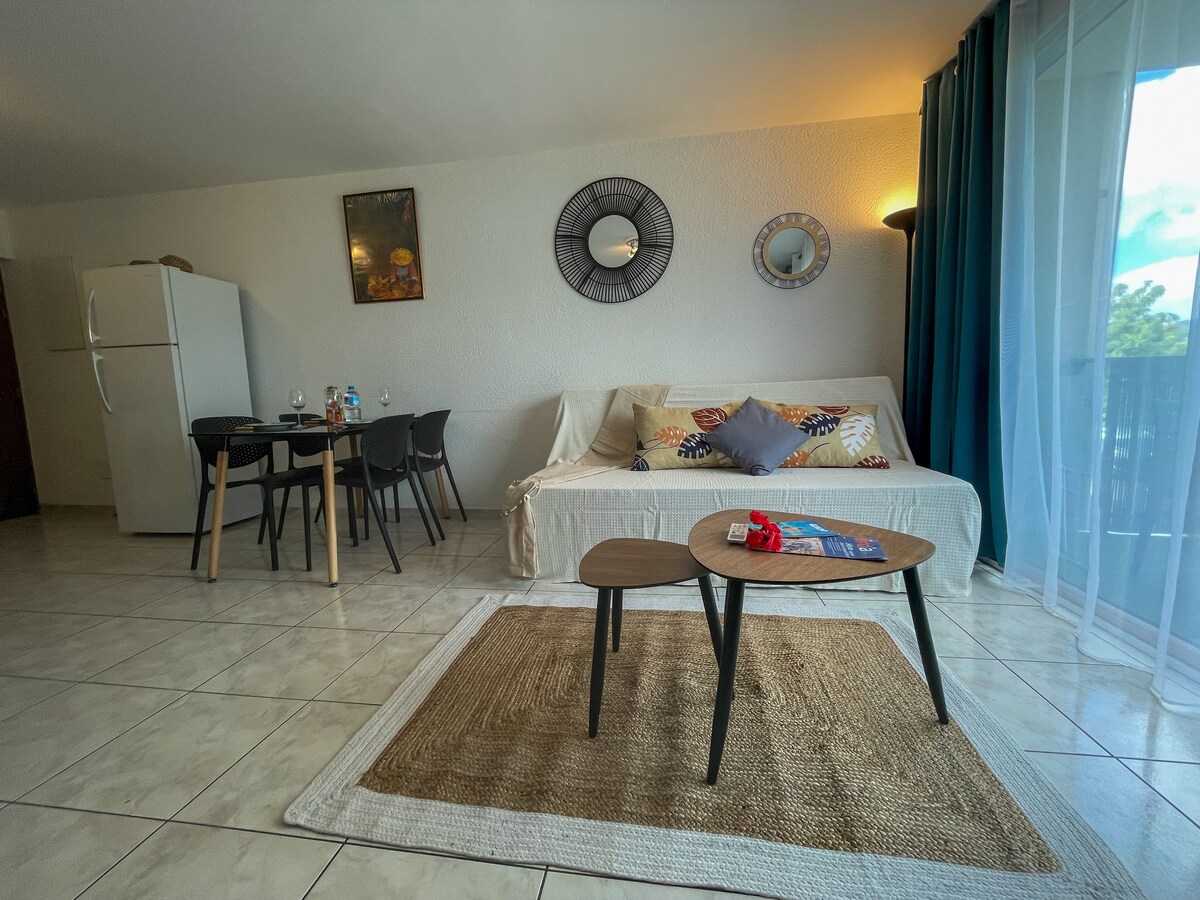 A spacious living area features a light-colored sofa bed adorned with decorative cushions. A round coffee table is positioned between two chairs. A full-size refrigerator is visible, along with a small dining table set for four. Natural light filters through sheer curtains.