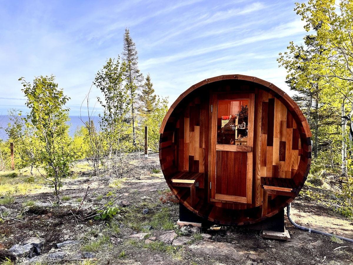 A panoramic barrel sauna is nestled among lush greenery, providing a serene escape. The wooden structure features a front door and benches inside, with Lake Superior visible in the background under a clear sky.