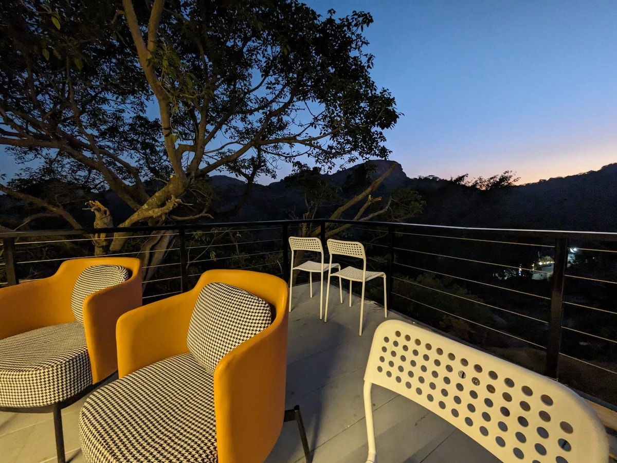 An outdoor seating area features two vibrant armchairs in orange and a checkered design beside white chairs. A railing provides a view of the valley and mountains, with the sky transitioning into twilight shades. The serene environment invites relaxation.