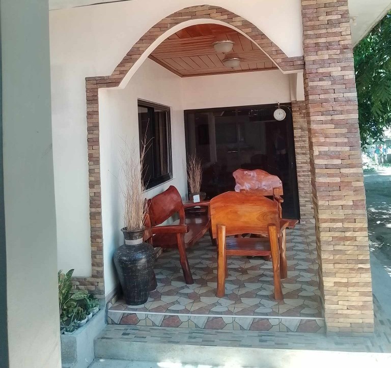 A covered entryway features traditional wooden furniture, including two chairs and a small table. Natural stone columns frame the entrance, and decorative plants can be observed at the base. Light-colored tiles create a welcoming path leading into the space.