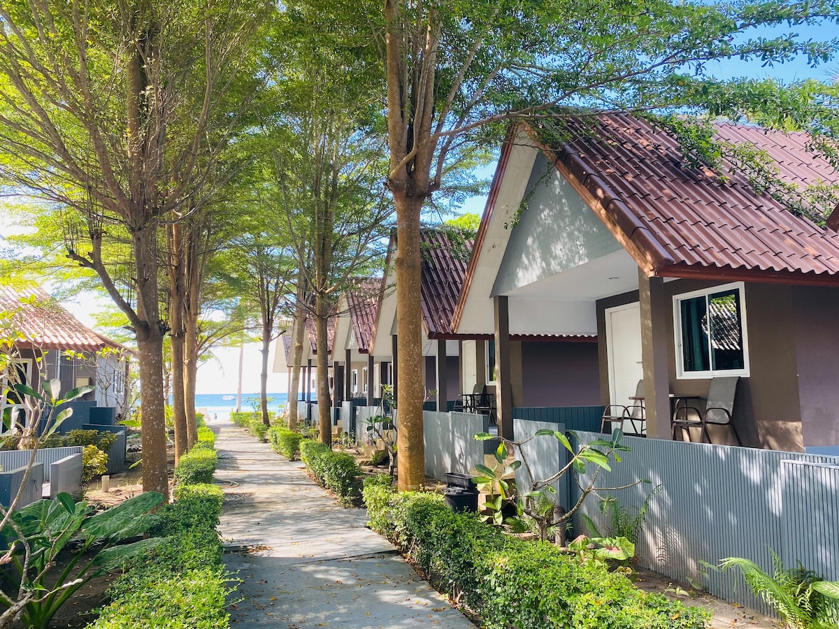 A pathway lined with trees leads to a row of Thai-style bungalows, featuring sloped roofs. The serene ocean is visible in the background, and lush greenery borders the path, providing a natural ambiance.