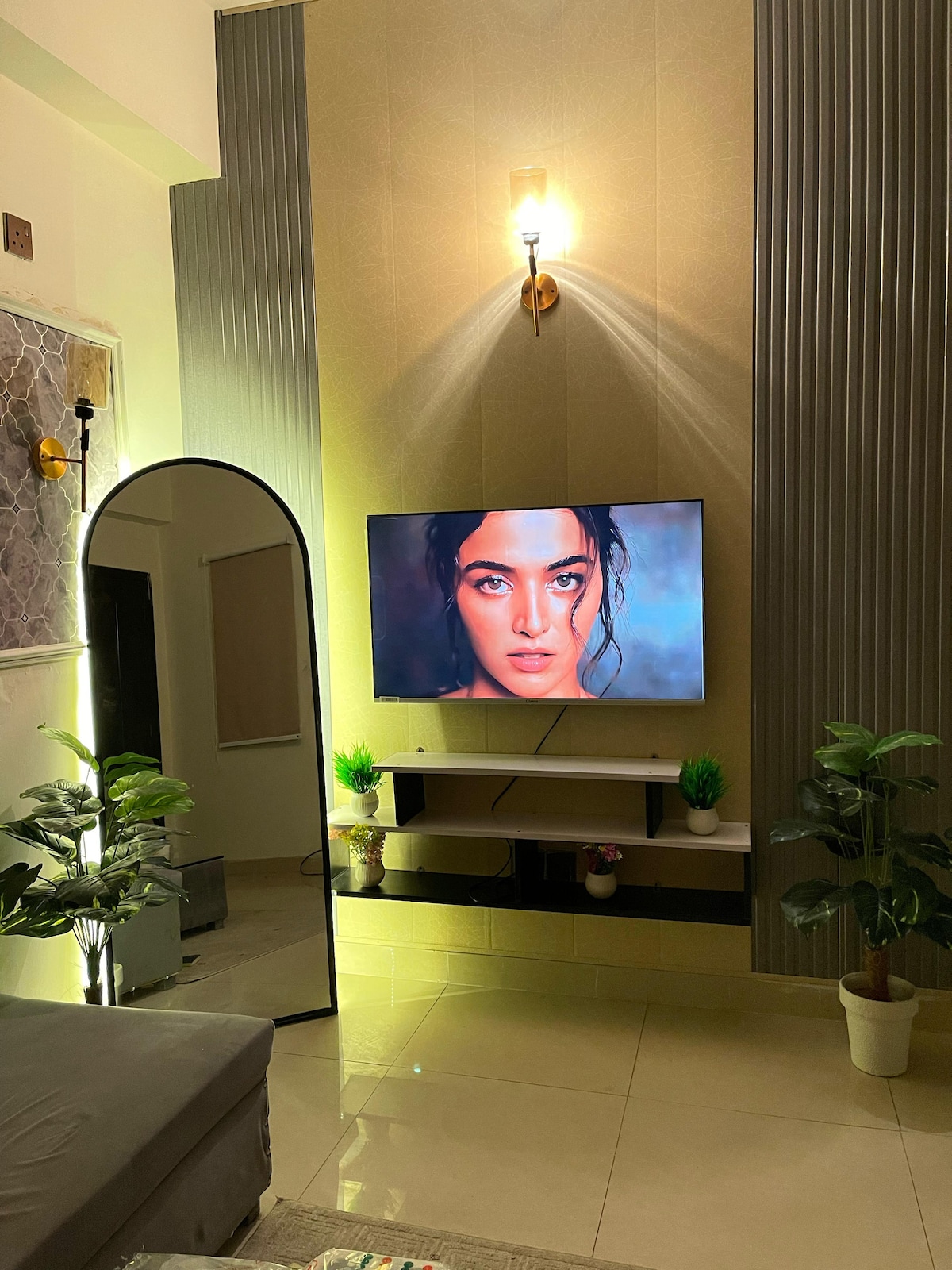 A modern lounge area is showcased with a large wall-mounted smart TV and a stylish mirror reflecting soft ambient lighting. Potted plants are positioned nearby, adding a touch of greenery to the clean, contemporary decor. The glossy floor enhances the overall spacious feel.