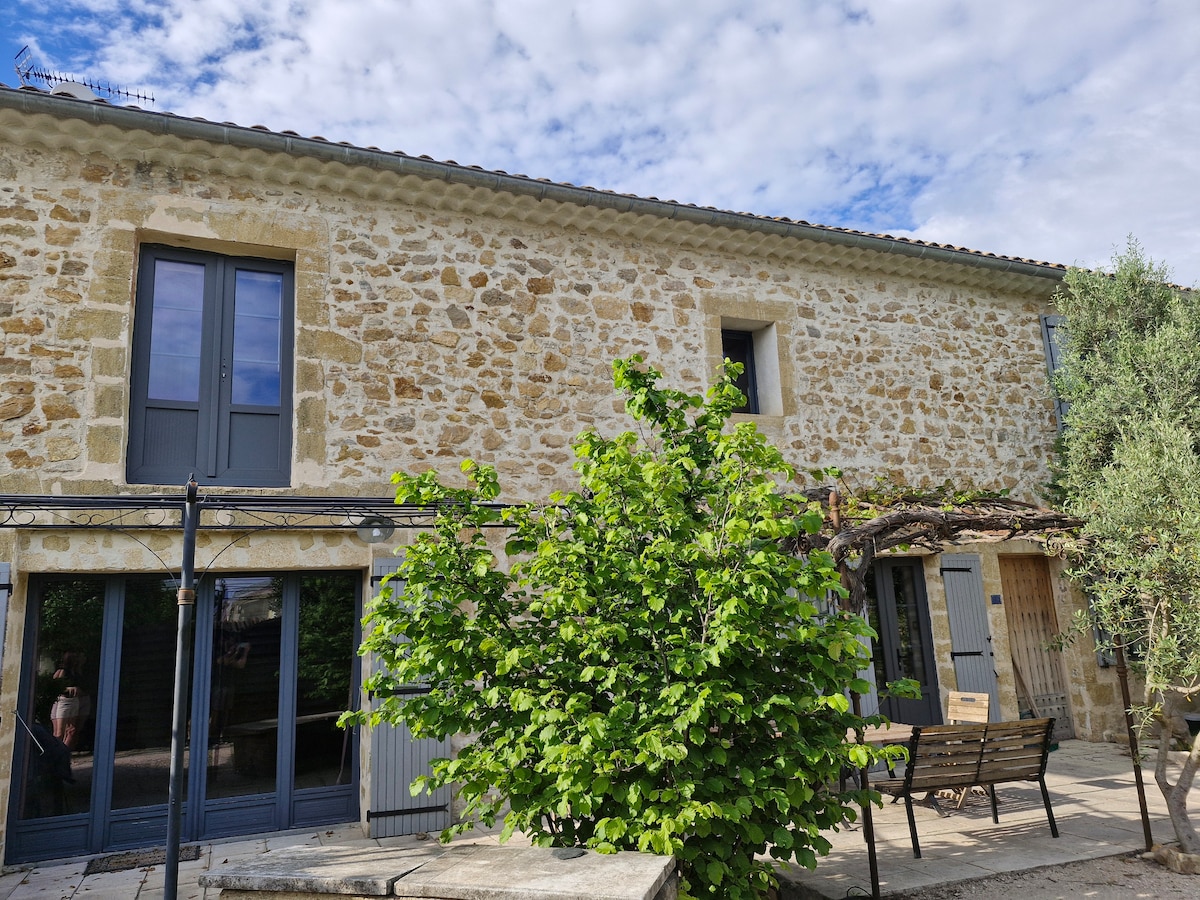 The exterior of the stone house is displayed, featuring large windows and a shaded outdoor area. A green tree provides lively contrast against the warm tones of the stone facade. The sky is partly cloudy, creating a serene atmosphere.