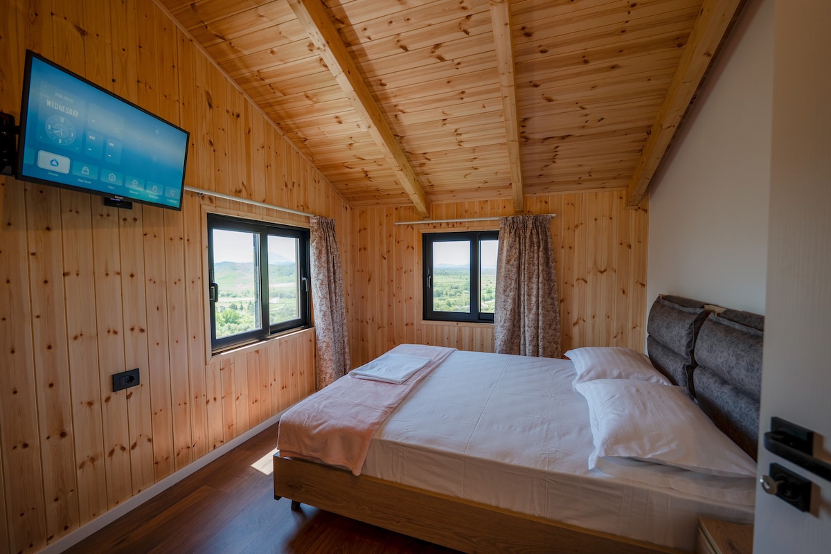 A cozy bedroom features a wooden ceiling and walls, creating a warm ambiance. A spacious bed is positioned against one wall, with white linens arranged neatly. Two large windows offer views of the surrounding landscape, and a television is mounted opposite the bed.