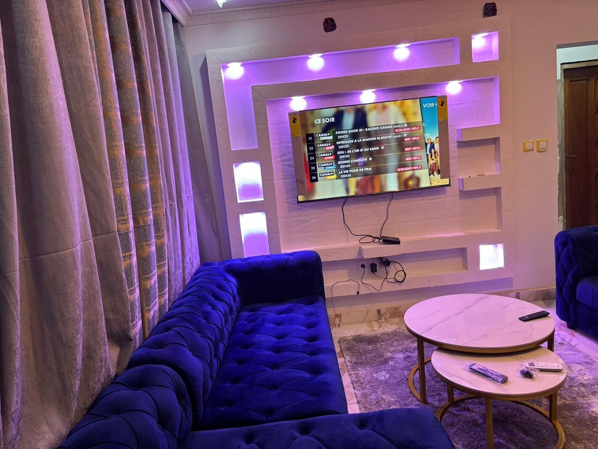 A modern living area is shown, featuring a plush blue sofa complemented by a circular coffee table. A flat-screen TV is mounted on the wall, with ambient lighting creating a soft glow. Elegant curtains frame the large window, enhancing the overall aesthetic.