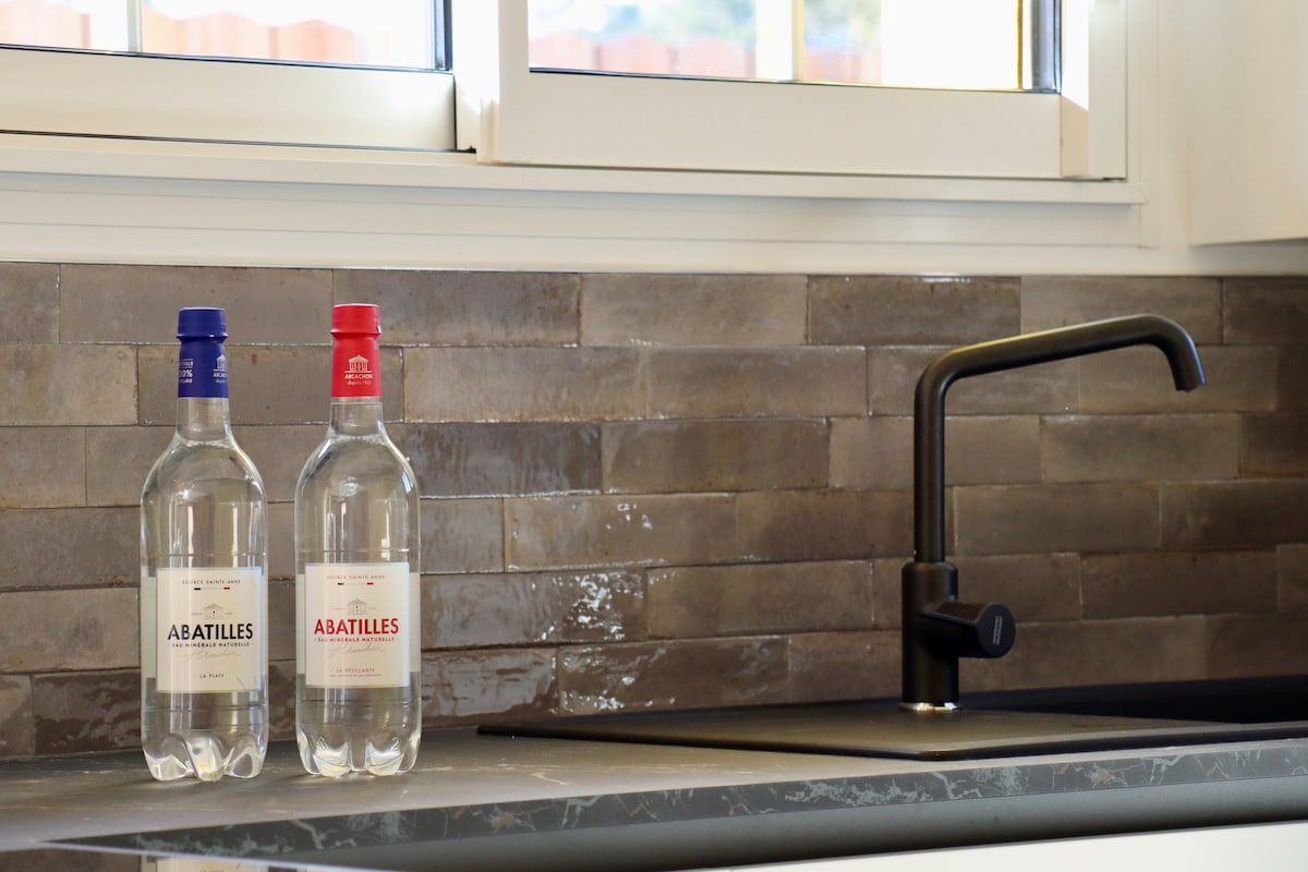 A kitchen countertop features two bottles of water, one with a red cap and the other with a blue cap, both labeled 'ABATILLES.' A sleek, black faucet is positioned next to the bottles, with a window providing natural light above.