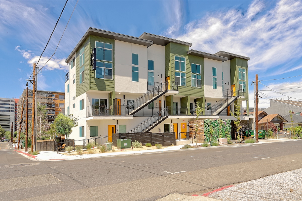 A modern multi-story building features a combination of green and neutral exteriors. Large windows provide ample natural light, and external staircases lead to different levels. Surrounding landscaping includes soft greenery, with a clear blue sky above enhancing the contemporary architecture.