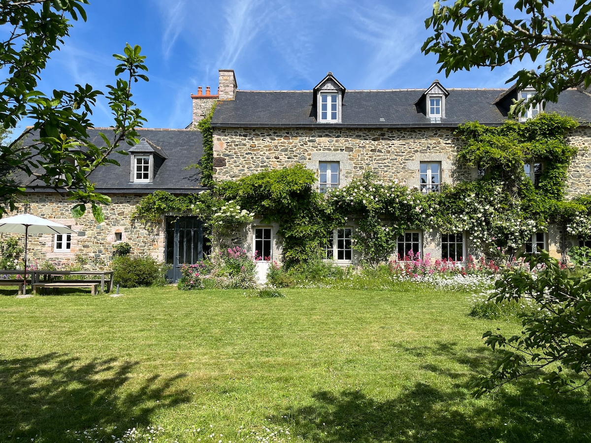 The stone house features a lush garden filled with colorful flowers and greenery. Windows are framed by climbing plants, and a spacious lawn offers an inviting outdoor space. A charming patio area with seating is visible, surrounded by a peaceful natural setting.