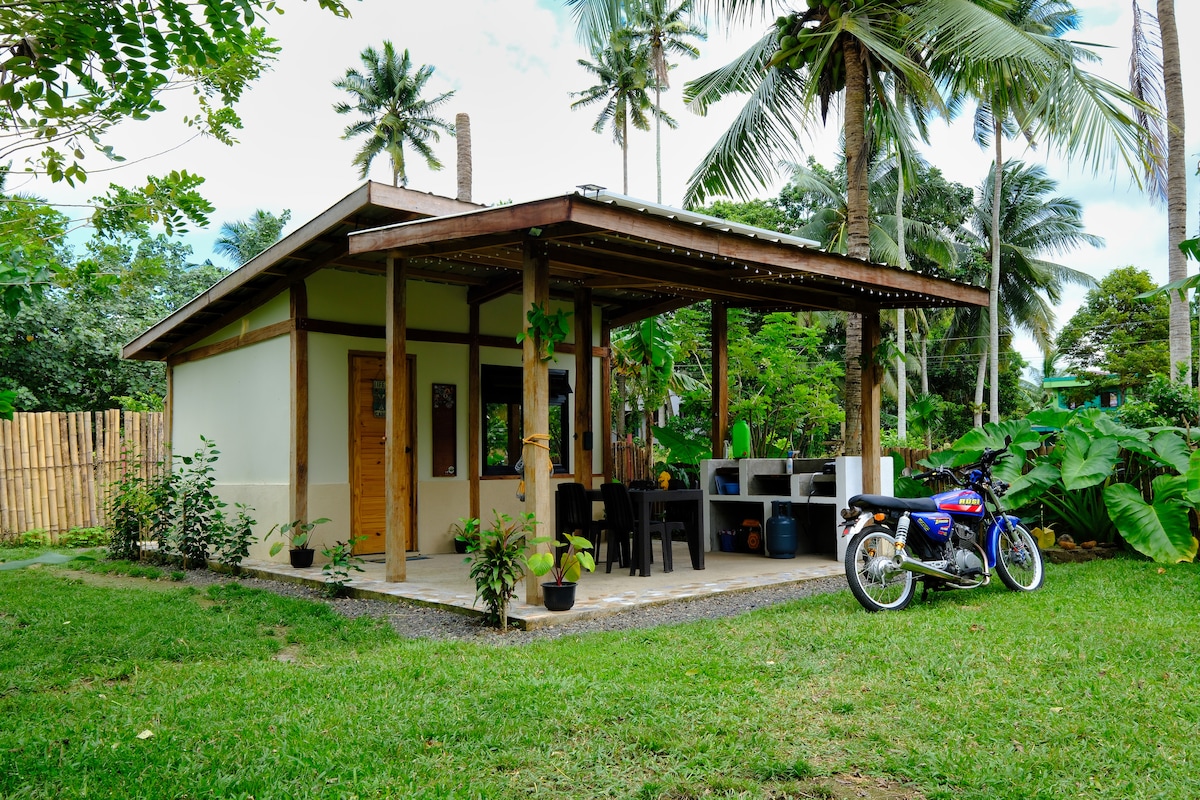 A simple, locally designed cabin features a spacious patio equipped with a cooking area and dining set. Lush greenery surrounds the structure, complemented by tall palm trees. A motorcycle is parked nearby, indicating options for exploration in the natural setting.