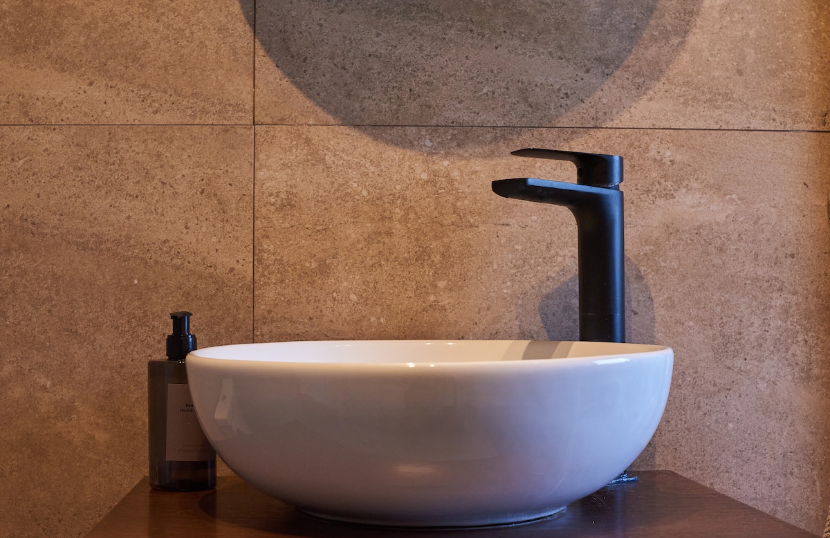 A sleek, modern basin is positioned atop a wooden countertop. A stylish black faucet extends over the bowl, complementing the minimalist design. A round mirror reflects the subtle textures of the warm stone wall behind, creating a harmonious and functional space.